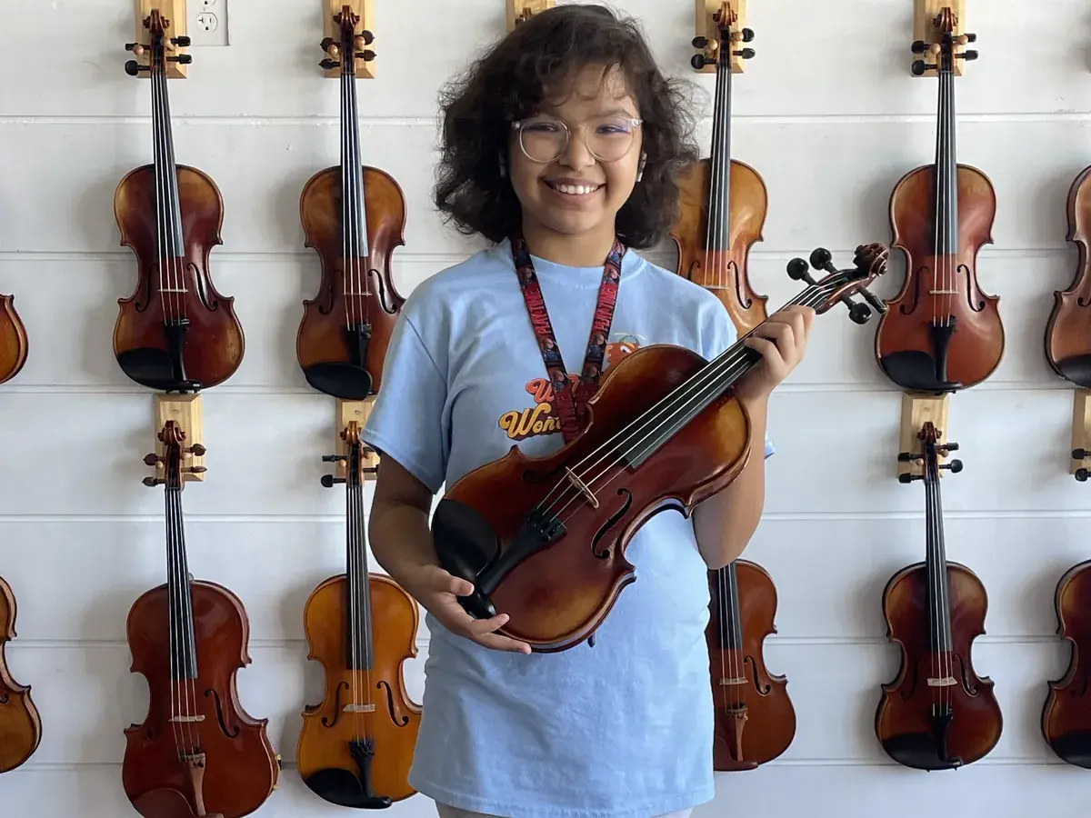 A smiling student holding a rental violin in front of the Bosky Strings violin wall